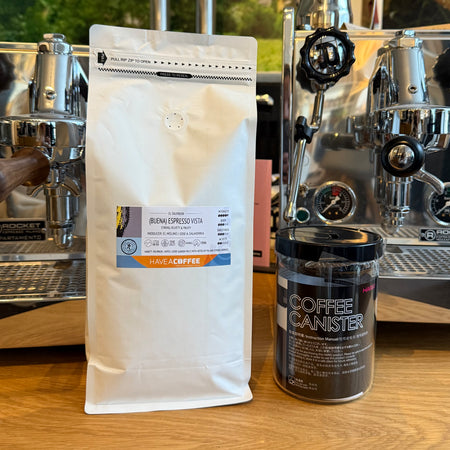 Pose med specialty coffee fra Have A Coffee og sort kaffeopbevaring placeret foran professionelle espressomaskiner i cafémiljø.