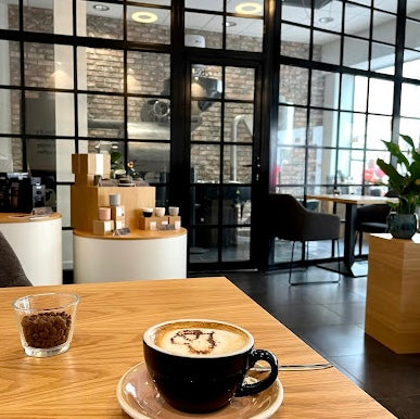 Cappuccino serveret i sort kop på træbord i moderne café med glasvægge, loungeområde og minimalistisk indretning i baggrunden.