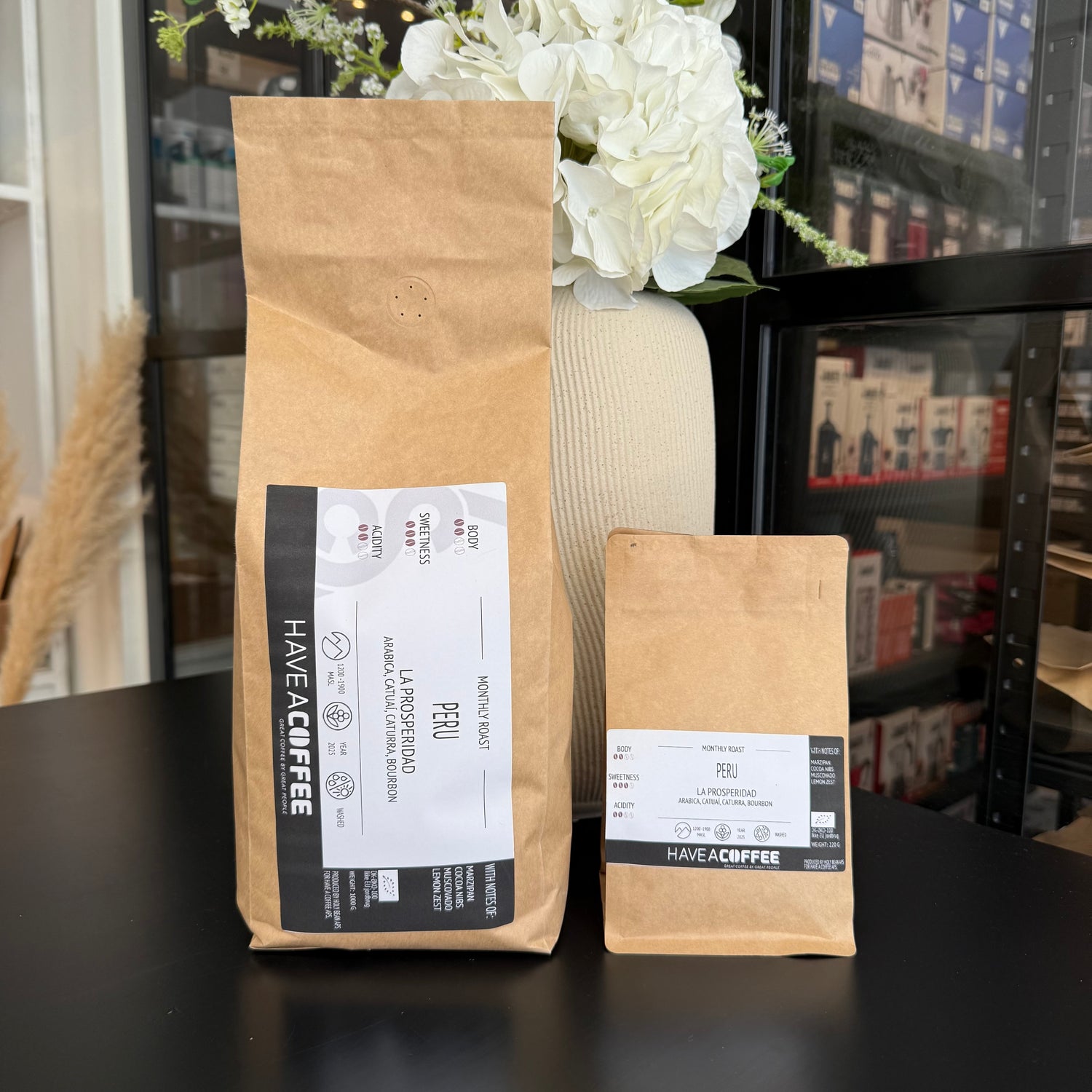 To poser Peru La Prosperidad specialty coffee fra Have A Coffee præsenteret foran hvid blomsterdekoration i moderne café- og showroommiljø.