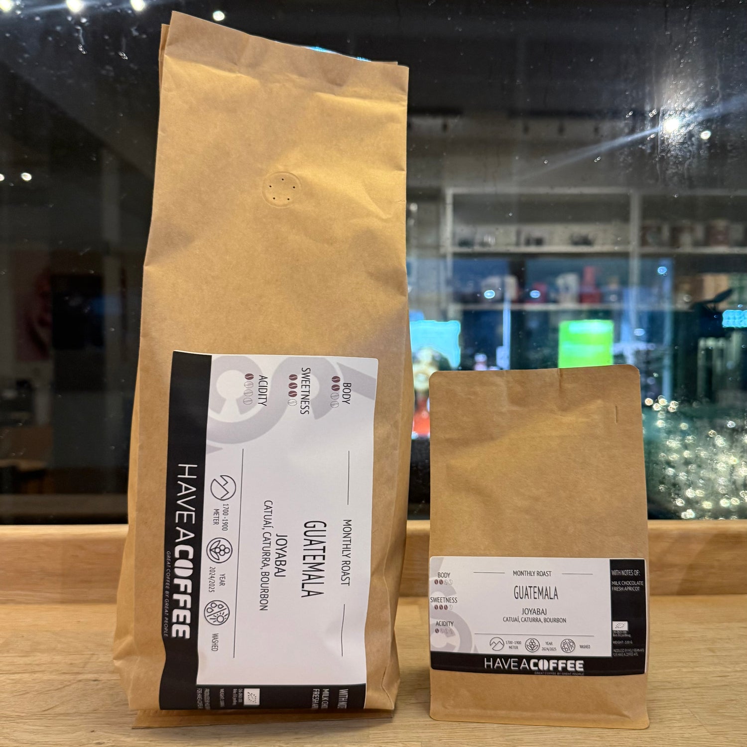 To poser Guatemala Joyabaj specialty coffee fra Have A Coffee placeret på træbord foran vindue med stemningsfuld cafébelysning i baggrunden.