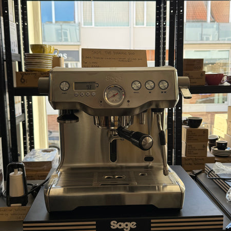 Professionel Sage espressomaskine i rustfrit stål udstillet i kaffebar med kopper og kaffetilbehør i baggrunden.