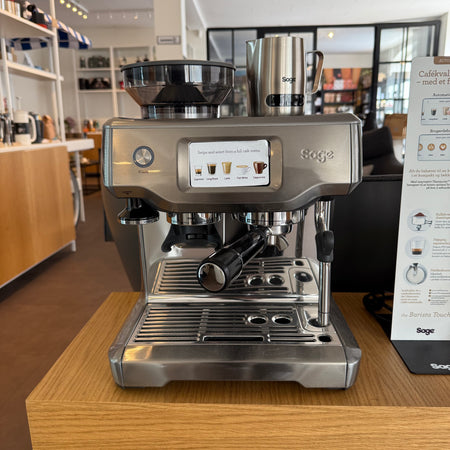 Moderne Sage Barista Touch espressomaskine med integreret touchskærm og mælkekande placeret på træbord i stilfuldt cafémiljø.